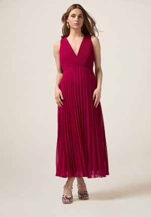 Largo vestido plisado en un vibrante magenta con un escote en V profundo y volantes en los hombros, acompañado de sandalias plateadas con tiras. Tela suave y fluida.