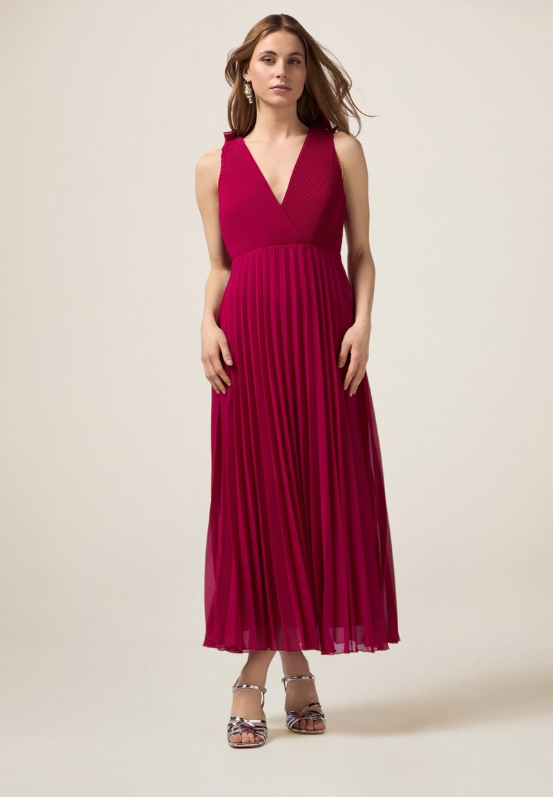Lange, geplooide jurk in levendig magenta met een diepe V-hals en schouderruffles, gecombineerd met zilveren sandaaltjes met bandjes. Glad, vloeiend materiaal.