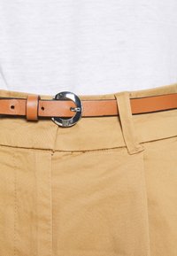 Pantalon en coton beige avec passants de ceinture apparents, muni d'une ceinture en cuir marron avec une boucle ronde argentée. Texture lisse, design simple.