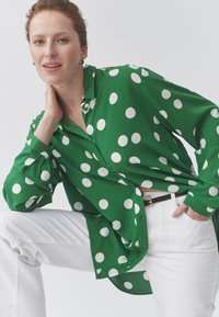 Žalia marškiniai su taškuotais apskritimais, ilgomis rankovėmis, su sagomis ir laisvo kirpimo, derinami su baltais džinsais ir juodu diržu.