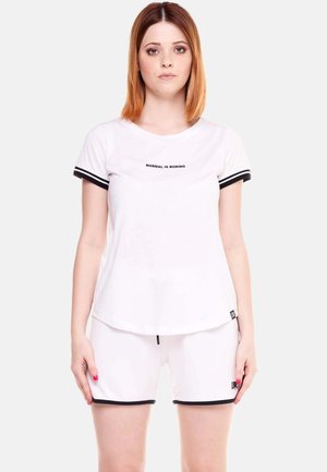 Donna con capelli lisci castano ramati fino alle spalle indossa una maglietta bianca con la scritta "NORMAL IS BORING" e pantaloncini bianchi abbinati con rifiniture nere.