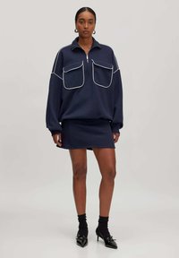 Marineblauwe oversized sweatshirt met witte piping en twee grote borstzakken, gecombineerd met een strakke marineblauwe mini-jurk en puntige zwarte hakken.