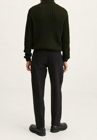 Khaki geribbelde gebreide coltrui gecombineerd met zwarte cargobroek met zijzakken en zwarte enkellaarzen, gezien van achteren.