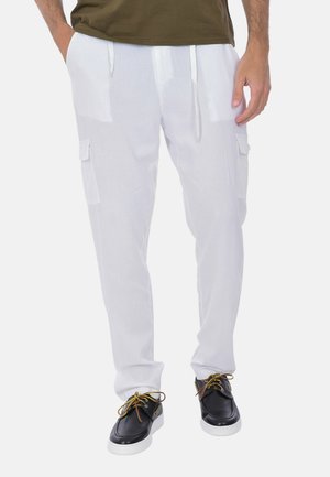 Uomo che indossa pantaloni cargo bianchi con tasche laterali, abbinati a scarpe stringate nere e bianche, in piedi con le mani nelle tasche.