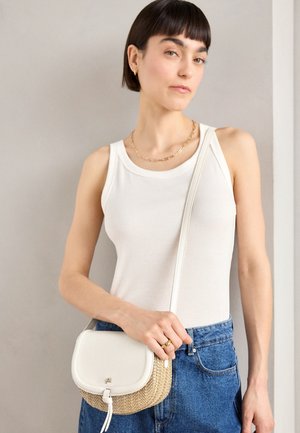 Vrouw draagt een witte mouwloze top en blauwe spijkerbroek, houdt een klein beige-wit crossbody tasje vast en staat tegen een effen achtergrond.