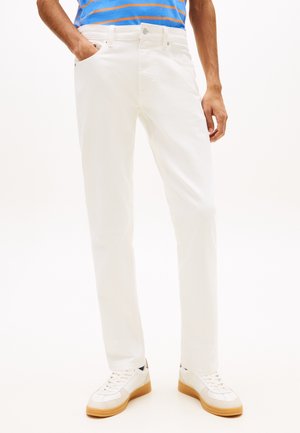 Fehér egyenes fazonú nadrágot, fehér, gumitalpú tornacipőt és kék-narancssárga csíkos inget viselő férfi, aki egyik kezét a zsebében tartva áll.