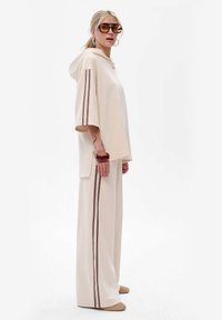Beżowa bluzka z kapturem i krótkimi rękawami, z bordowymi paskami po bokach, zestawiona z szerokimi beżowymi spodniami o luźnym kroju i fakturowanej tkaninie.