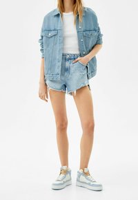 Světle modrá džínová bunda se dvěma náprsními kapsami, spárovaná s roztrhanými světle modrými šortkami a bílými teniskami s modrými akcenty a krémovými tkaničkami.