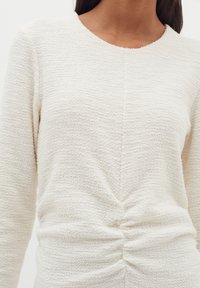 Krämfärgad stickad topp med texturerad tyg, rund halsringning och rynkdetalj vid midjan, som ger en åtsittande silhuett.