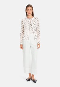 Weißer Cardigan mit braunen Punkten, runder Ausschnitt und gewellten Kanten, kombiniert mit hochtaillierten weißen Hosen und schwarzen Schuhen.