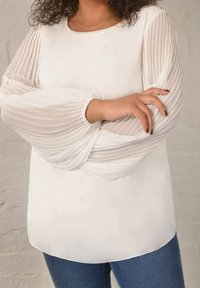 White long-sleeve top with pleated sheer sleeves, round neckline, and a smooth, fitted body. Paired with blue denim jeans.