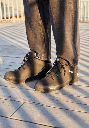 Osoba nosząca czarne buty trekkingowe Timberland i ciemnoszare spodnie stojąca na oświetlonym słońcem drewnianym tarasie z liniami cienia.