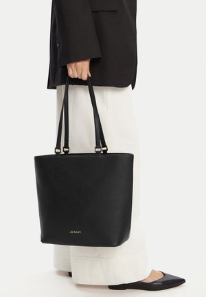 Persona che tiene in mano una borsa tote in pelle nera con doppi manici, indossando pantaloni larghi bianchi e ballerine nere a punta.