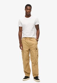 Pantalons cargo beiges avec poches latérales, taille élastique et coupe décontractée, portés avec un t-shirt blanc uni et des baskets noires.