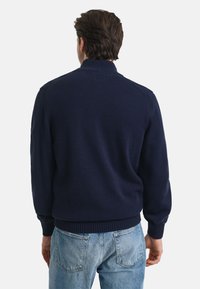 Temno modra pletena pulover s visokim ovratnikom, rebrastimi manšetami in robom, ki se ponaša z mehkobo in sproščenim krojem, prikazan od zadaj.