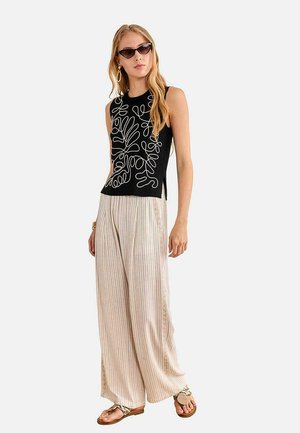 Mujer con top sin mangas negro con líneas abstractas blancas, pantalones beige de pierna ancha, sandalias marrones, gafas de sol estilo ojo de gato y pendientes de aro.