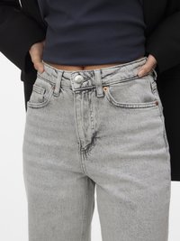 Persoon draagt een lichtgrijze hoge taille jeans met koperen klinknagels en een zwart cropped topje, handen in de voorkant zakken gestopt.