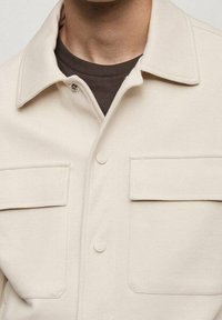Beige jakke med krave og frontlommer, båret over en mørkebrun crew neck trøje, der kun viser øverste del af brystet og halsen.