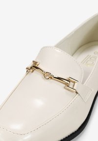 Mocasín de charol crema con acento de cadena dorada y suela de goma negra. Presenta una punta redondeada y detalles de costura a lo largo de los costados.