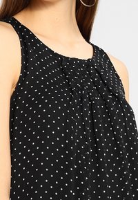 Top senza maniche nero con pois bianchi, caratterizzato da un colletto arricciato e un tessuto morbido e testurizzato, progettato per una vestibilità fluida.