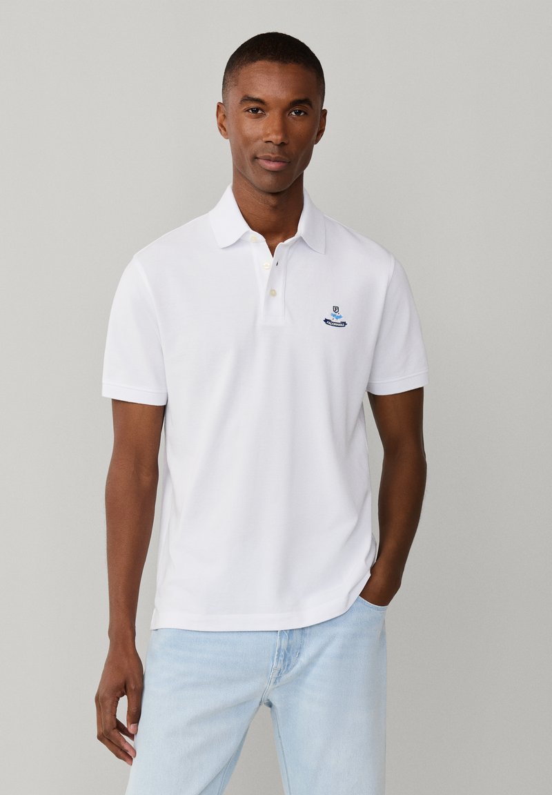 Man staat, draagt een wit poloshirt met korte mouwen en een klein blauw logo op de linkerborst, en lichtblauwe spijkerbroek, neutrale achtergrond.