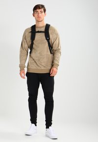 Beige kamuflasje sweatshirt, svarte bukser og hvite sneakers. Har en frontstropp og ribbede mansjetter. Enkelt og funksjonelt design.