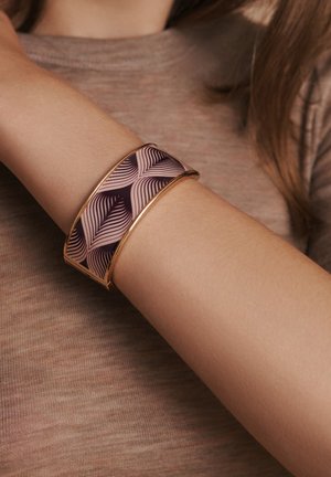 Gold-tone cuff bracelet featuring a glossy purple and pink wave pattern, worn on a light skin tone arm against a textured taupe background.