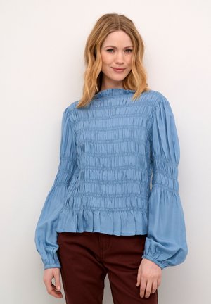Blusa azzurro chiaro con texture arricciata, colletto rotondo e maniche lunghe. Presenta un orlo volante e un tessuto liscio. Abbinata a pantaloni bordeaux.