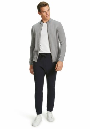 Gilet zippé gris sur un t-shirt blanc, accompagné de joggings navy et de baskets blanches. Mettez l'accent sur les matériaux et la coupe ajustée.