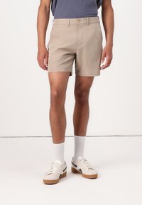 Shorts beiges à l'avant plat, avec deux poches latérales et une fermeture par bouton. Portés avec des chaussettes blanches et des baskets crème avec des accents noirs.