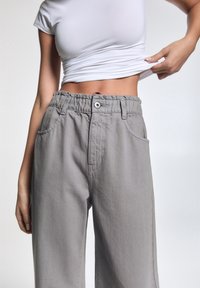Pantalon décontracté gris clair, ample, avec une taille élastique, deux poches avant et un bouton de fermeture. Texture lisse et douce, design casual.