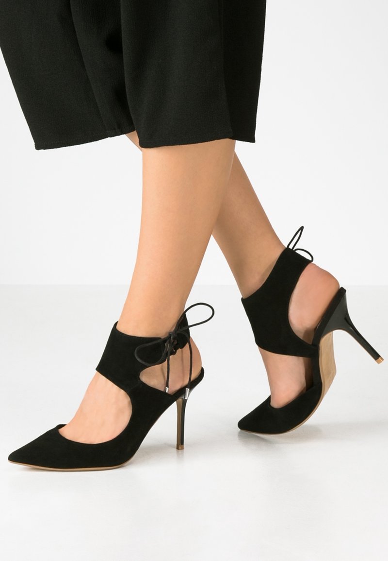 Jambes portant des escarpins noirs à bouts pointus avec découpes à la cheville et lacets, associés à un pantalon noir coupé au-dessus de la cheville.