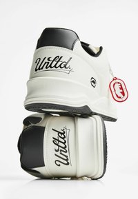 Valkoiset tennarit mustilla yksityiskohdilla, "Unltd." logo kantapäässä, kerrosrakenteinen korko ja punainen avaimenperä, jossa on karhun siluetti.