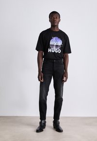 T-shirt en coton noir avec un graphique de palmier en dégradé et le texte "HUGO". Porté avec un jean en denim foncé et des bottines noires.