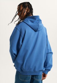 Sudadera azul con capucha y un ajuste relajado, que presenta una textura suave, puños acanalados y una pequeña etiqueta de logo en el dobladillo. Se muestra vista posterior.