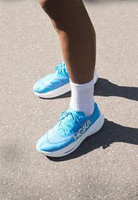 Blue athletic shoes with a textured mesh upper, white sole, and prominent "HOKA" branding, paired with white ankle socks on asphalt.