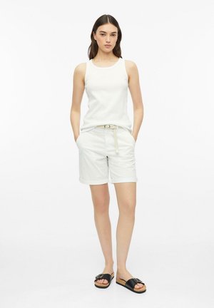 Vrouw staand met handen in de zakken, gekleed in een witte mouwloze top, witte short met riem en zwarte instapsandalen tegen een witte achtergrond.