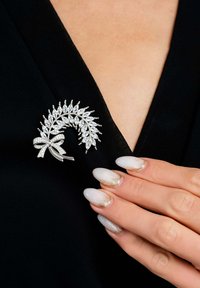 Spilla in tonalità argento con design a foglia, adornata con strass trasparenti. Presenta un accento a forma di fiocco, posizionata su uno sfondo di tessuto nero.