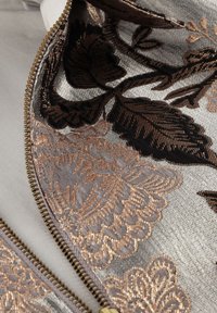 Geborduurd fabric met zilveren, zwarte en koperen bloemmotieven. Inclusief een gouden rits langs de rand, met een soepele textuur.