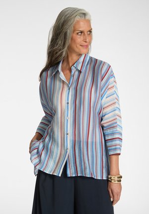 Kobieta z siwymi włosami ubrana w luźną, długą koszulę zapinaną na guziki z pionowymi niebieskimi, czerwonymi i beżowymi paskami oraz ciemne spodnie, patrząca w prawo.