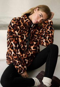 Veste zippée en peluche avec un motif léopard dans des teintes orange et noire, dotée d'un col haut et d'épaules tombantes. Associée à des leggings noirs.