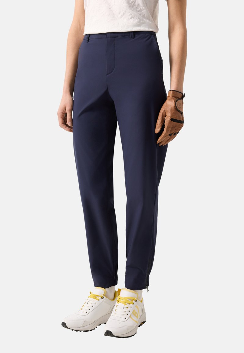 Osoba nosząca granatowe spodnie o prostych nogawkach, białe trampki z żółtymi akcentami oraz brązową skórzaną rękawiczkę na prawej dłoni.