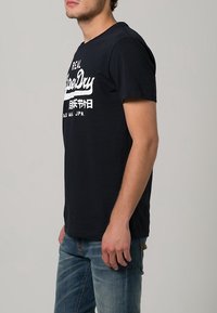 Černé bavlněné tričko s krátkým rukávem a bílým nápisem. Nosené s modrými džíny, které mají klasický střih a minimalistický design.