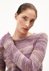 Frau mit zurückgegelten Haaren, die einen lila und beigen gestreiften Strickpullover trägt, blickt mit einem neutralen Ausdruck in die Kamera, Hand nahe der Schulter.