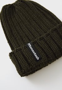 Olive green knit beanie with a ribbed texture, featuring a folded cuff and a black brand label sewn into the front.