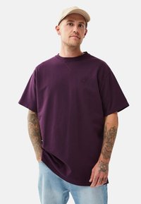 Mann mit tätowierten Armen, der eine beige Kappe, ein übergroßes dunkelviolettes T-Shirt und eine hellblaue Jeans trägt und vor einem schlichten Hintergrund steht.