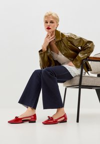 Ballerine in vernice rossa con design a taglio e dettaglio a fiocco. La modella indossa una giacca verde lucida e jeans svasati scuri, seduta su una sedia.