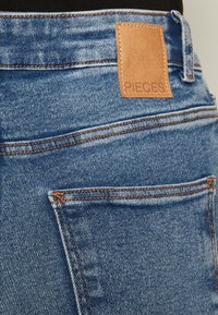 Jeanși din denim cu o culoare bleu deschis și un finisaj texturat, eticheta din piele maro cu inscripția "PIECES" și accente de cusătură portocalie.