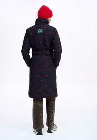 Čierna prešívaná bunda s červenými bodovými akcentmi, vysokým golierom, opásaným pásom a zelenou nášivkou na zadnej strane, spojená s nohavicami s leopardím vzorom.