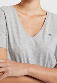 Světle šedé tričko s výstřihem do V, které má na levé straně hrudi malé logo. Paže jsou zkřížené před tělem.